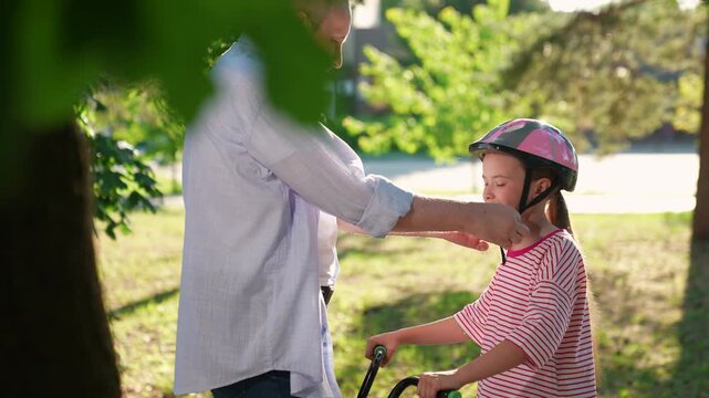 Dad puts protective helmet on little daughter rides bike outdoors in summer. Kid girl cyclists with papa in city park. Family weekend. Father teaches daughter to ride bike park. Happy sporty family