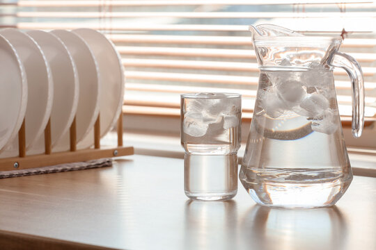 Refreshing cold water pitcher and glass with ice cubes on kitchen countertop by window blinds, sunlight streams through window blinds in the background, creating reflections on the polished surface