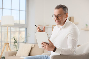 Fototapeta premium Smiling mature man using digital tablet and stylus pen for online learning or business communication at home, he is casually dressed in a white shirt and glasses, seated comfortably