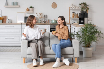Obraz premium Two young women relaxing on sofa with coffee mugs in modern kitchen living room interior, they are holding coffee mugs and engaged in a pleasant conversation, smiling and enjoying each others company