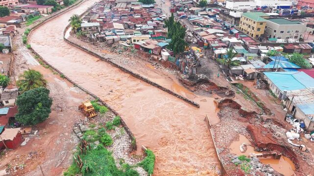 Rising aerial view, floodwater rushes through a storm drain in a densely populated low-income community during severe flooding in Accra, Ghana