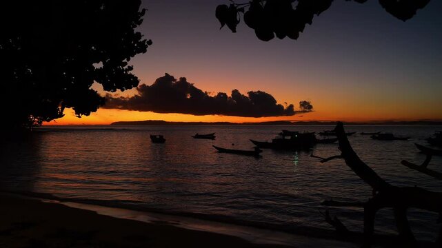 SILHOUETTE: Moored boats float in calm water as the last orange glow fades behind a dramatic cloud bank. Seascape framed by shadowy coastal trees and quiet twilight mood in the Mentawai Islands.