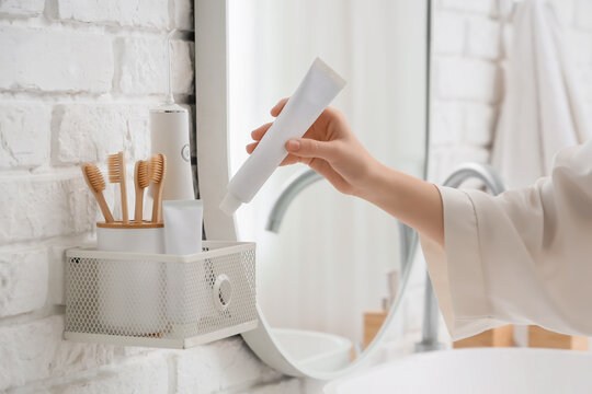 Womans hand holding a blank white toothpaste tube in a modern bathroom with eco-friendly bamboo toothbrushes and oral irrigator