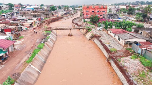 Aerial view, Muddy floodwater rushes through a concrete storm drainage channel in a densely populated residential neighborhood in Accra, Ghana