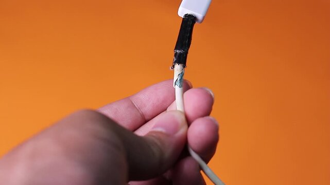 Close-up video frame showing a hand holding a bending, broken white charging cable with insulation peeling away from the wire joint.