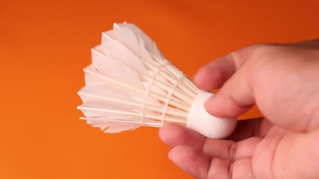 Side view video frame of a hand gently holding a white feather shuttlecock horizontally over a solid bright orange background.