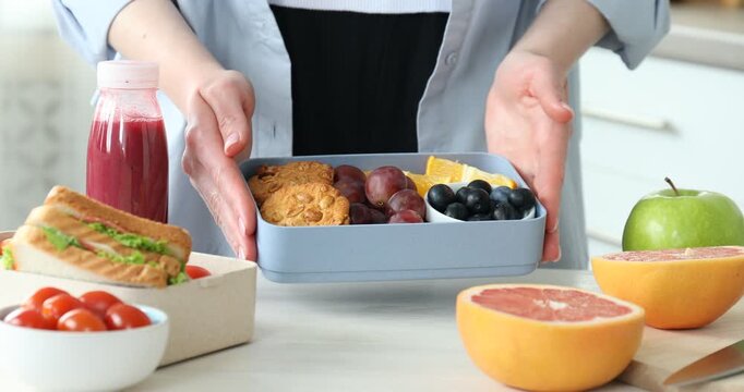 Woman putting fresh cookies into lunch box and showing it at light wooden table indoors, closeup