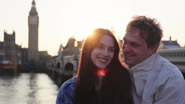 Love couple embraces tenderly near Westminster Bridge in London with Big Ben silhouette in golden sunset light. Young man and woman share intimate moment by River Thames during romantic holiday.