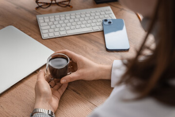 Fototapeta premium Young female doctor with cup of coffee, mobile phone and laptop sitting at table in hospital, closeup