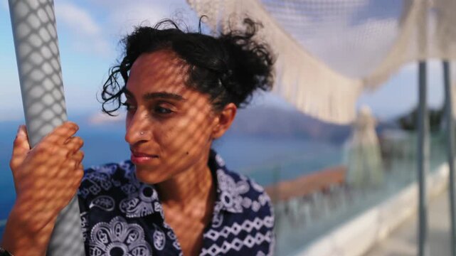 Beautiful woman with curly dark hair stands on a seaside terrace in Santorini Greece during golden hour. Mesh canopy creates diamond shadow patterns on her face as she gazes at the sea view.