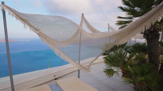 Woven macrame hammock with fringe sways gently in the breeze on a luxury terrace in Santorini Greece. Palm trees frame the stunning blue Aegean Sea and caldera view under cloudy skies.
