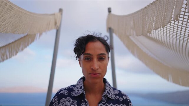 Young Indian woman in navy paisley outfit stands between two macrame hammocks on luxury terrace overlooking the Aegean Sea in Santorini Greece during cloudy evening with mountains visible in distance.