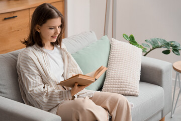 Obraz premium Beautiful young woman reading book on cozy sofa with soft pillows in living room