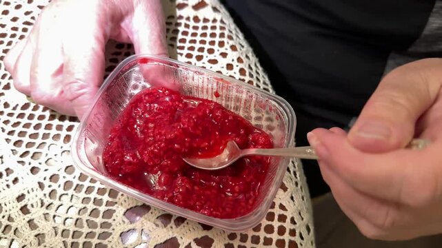 Frozen raspberries in plastic container stirred by woman with spoon close-up showing bright red icy texture on lace tablecloth hand