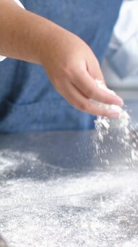 Vertical video: Reaching chef in blue apron sprinkling flour on steel top, metal bowl for dough