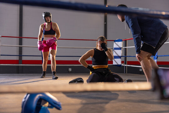 Female boxers and coach tending seated boxer holding face on ring canvas with blue gloves nearby