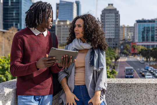 Diverse friends discussing tablet, holding backpack and leaning on concrete parapet over busy road