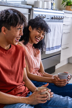 Diverse couple sitting on kitchen floor holding ceramic mugs and laughing after sharing coffee