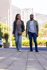 Fototapeta premium African American couple walking city plaza on paved path, woman wearing plaid overshirt and tan bag