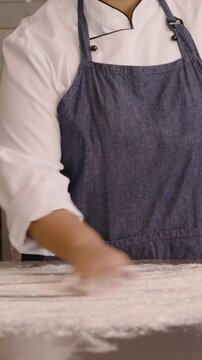 Vertical video: Reaching, chef spreading flour on metal counter for dough prep, wearing denim apron