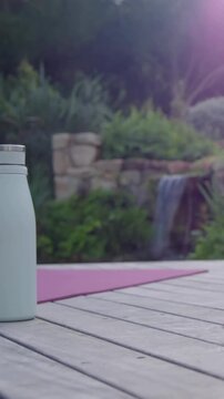 Vertical video: Camera panning, bringing water bottle onto wooden deck near pink yoga mat for yoga
