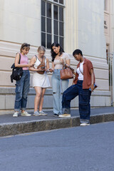 Fototapeta premium Diverse female friends standing on stone curb, checking smartphones and holding takeaway coffee cup