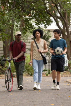African American males walking in park pushing bike with coffee, teenage male holding soccer ball