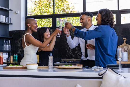 Diverse friends in party attire raising champagne flutes and clinking at kitchen island with pizzas