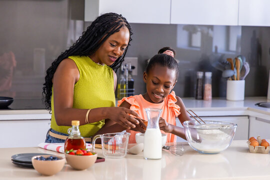 African mother guiding youth daughter, cracking egg into small white bowl at modern kitchen island