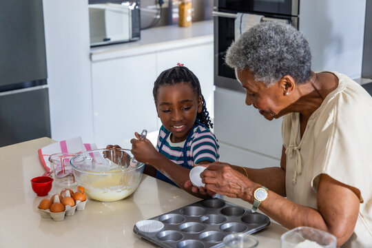 African American Senior woman and Child girl whisking batter at kitchen island, lining muffin tin
