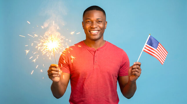 Black young adult man holding lit sparkler in left hand and small American flag in right hand on soft blue background, patriotic 4th of July portrait celebrating Independence Day, freedom and national