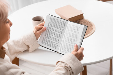 Naklejka premium Beautiful senior woman reading book at table in kitchen