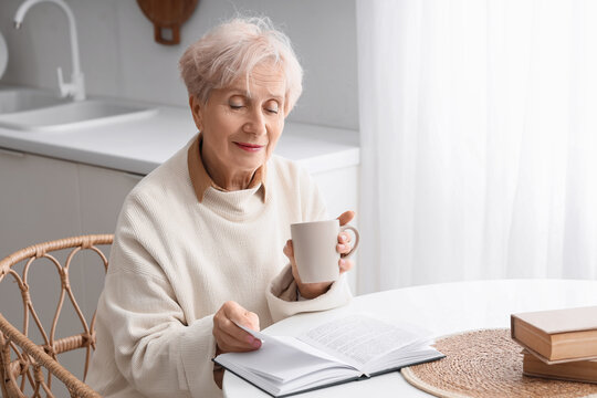 Beautiful senior woman with tea cup reading book at table in kitchen