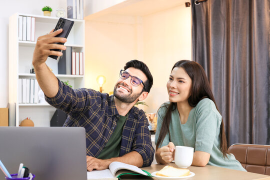 Business couple working, communicating and selfie while sitting at the home office desk together. Cheerful couple, calm asian woman and caucasian man selfi working at home with laptop together.