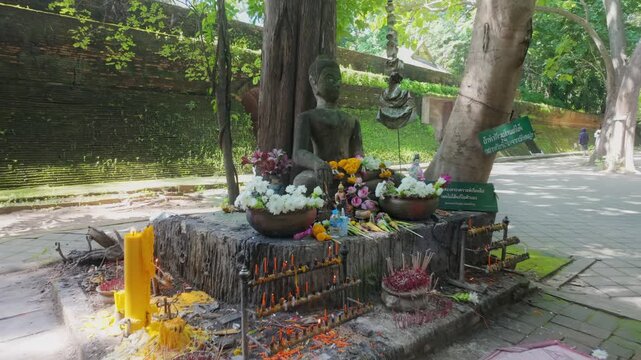 A weathered Buddha statue adorned with marigold garlands sits peacefully in a lush garden at Wat Umong, Chiang Mai, Thailand. The ancient temple grounds offer a serene atmosphere