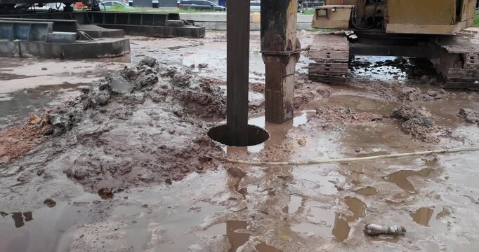 Industrial auger bores into a muddy, waterlogged construction site, pulling up wet soil and leaving heaps around the hole.