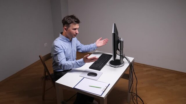 Young man sitting in an office, gesturing with frustration or confusion at a monitor during a video conference, indicating technical issues or misunderstanding a situation while working remotely
