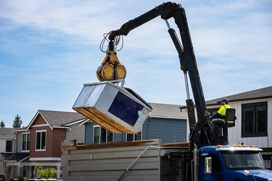 Construction dumpster being emptied by knuckle boom arm on specialized grapple truck, operator in control seat, new residential housing development
