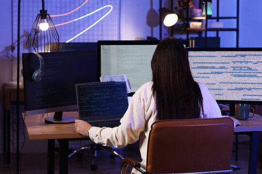 Young female programmer working with laptop in modern office at night, back view