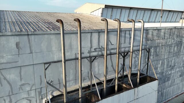 A drone shot showing a row of industrial metal exhaust pipes on the exterior wall of a factory building, with a corrugated roof in the background.