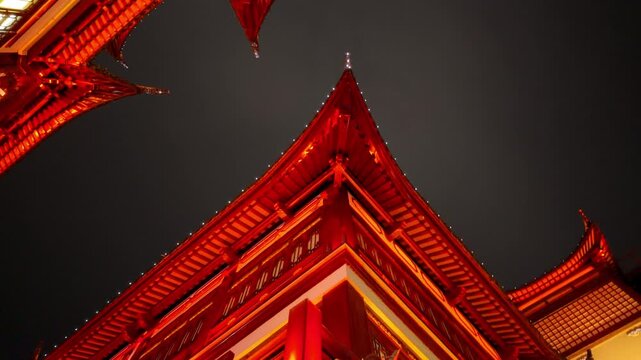 Traditional red-lit temple roofs illuminated at night against a dark sky, upward view