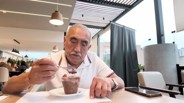 Side angle footage of a senior man eating chocolate mousse in a cafe. Retirement lifestyle, leisure, active aging and authentic everyday moments.