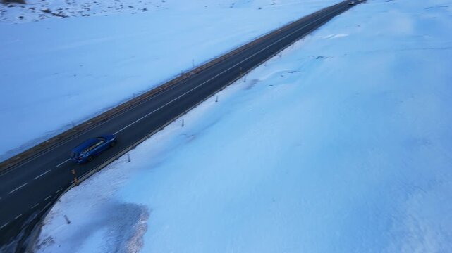 An aerial view captures a blue car driving on the Julier Pass road, surrounded by a vast winter landscape covered in snow and ice.