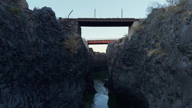 Rugged volcanic canyon in La Payunia Reserve, navigating directly underneath a striking orange bridge at sunrise. Slow, forward aerial glide.