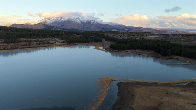 Milky light blue waters of Laguna La Zeta, framing lush shoreline reeds and snowy peaks at dawn. Slow, right tracking aerial.