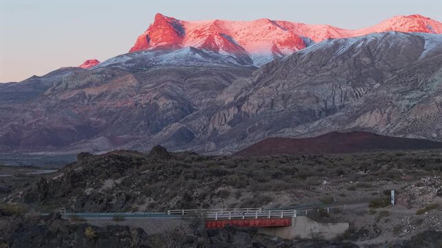 Snow-covered Andes dominate La Payunia Provincial Reserve, Malarg&uuml;e, Mendoza, Argentina, above volcanic formations, arid shrub steppe and a bridge crossing rugged terrain, slow lateral slide right