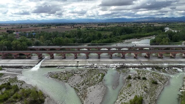 Parallel road and railway bridges cross the Trebbia River near Rottofreno, Piacenza, Emilia-Romagna, Italy, braided channels and gravel bars, with vehicle traffic visible on the roadway, slow push in