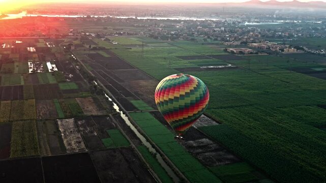 Aerial view of a multi-colored patterned hot air balloon gliding low over a patchwork of lush green agricultural fields and irrigation canals on Luxor's West Bank, with other balloons at sunrise.