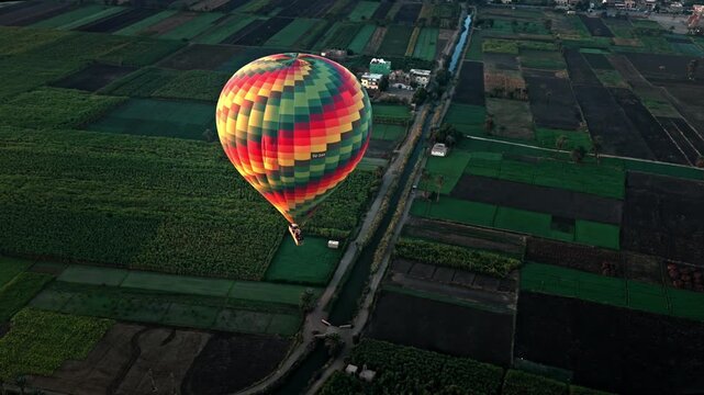 Aerial view of a multi-colored patterned hot air balloon gliding low over a patchwork of lush green agricultural fields and irrigation canals on Luxor's West Bank, with other balloons at sunrise.