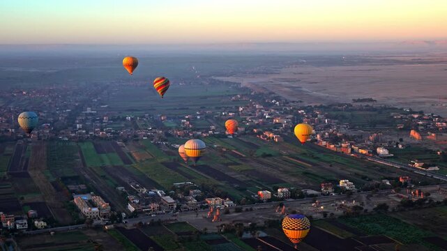 A high-angle aerial view capturing several colorful hot air balloons drifting over the green agricultural fields and archaeological ruins of Luxor's West Bank during a hazy golden sunrise.